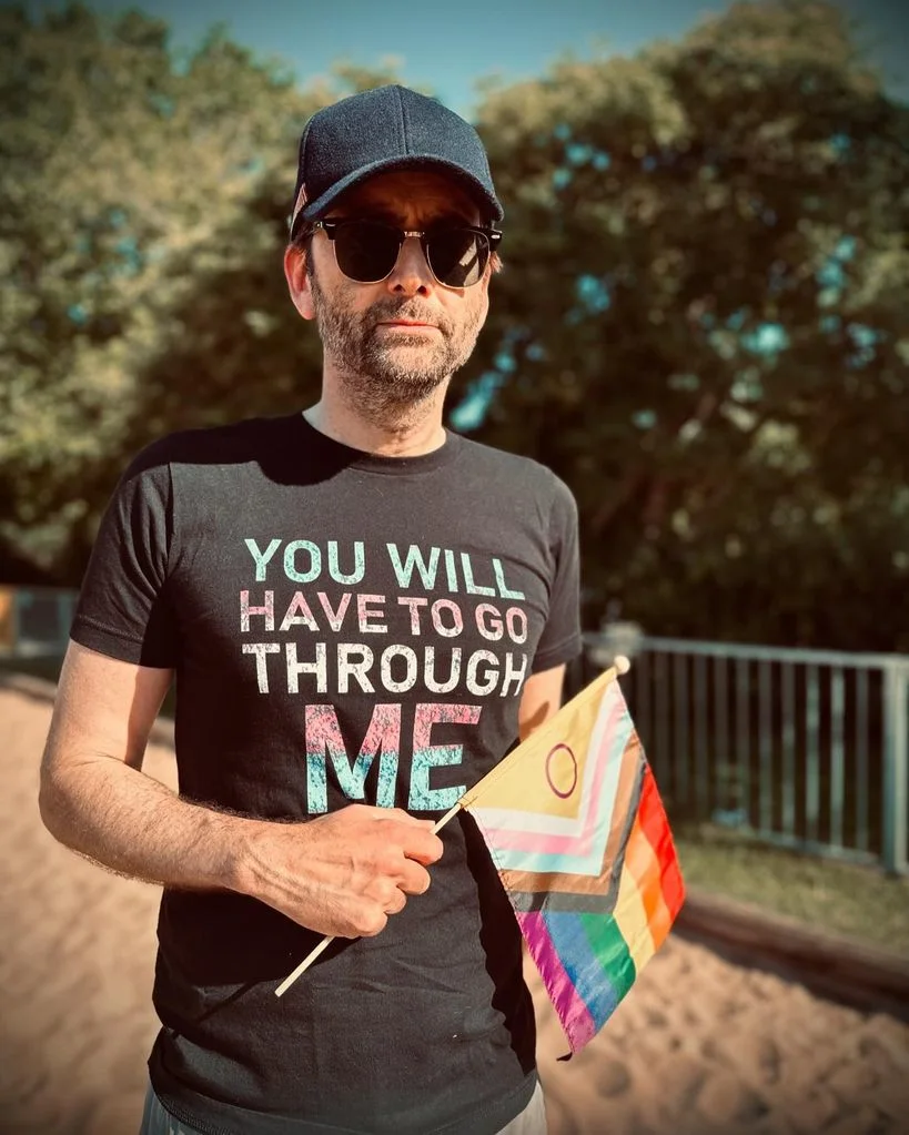 David Tennant. Lleva una bandera LGTBIAQ+, y una camiseta negra en la que pone "Tendrás que pasar por encima de mi". El mensaje lleva los colores de la bandera trans.